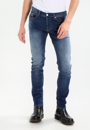 Jean bleu slim porté par une personne debout, associé à des bottines en cuir noires et une chemise blanche.