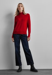 Roter Rollkragenpullover aus weicher Stoffqualität, kombiniert mit dunkelblauen weit geschnittenen Jeans und schwarzen Knöchelstiefeln, vor einem grauen Hintergrund stehend.