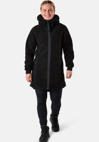 Schwarze Fleecejacke mit Kapuze, mit einem Reißverschluss vorne, seitlichen Taschen und gerippten Bündchen. Getragen über schwarzen Leggings und Stiefeln.