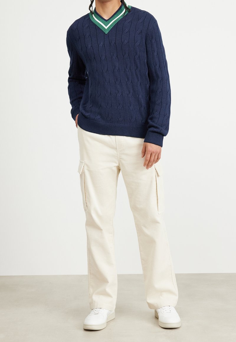 Homme portant un pull bleu marine à col en V tricoté en torsades et un pantalon cargo écru avec des baskets blanches, debout sur un fond uni.