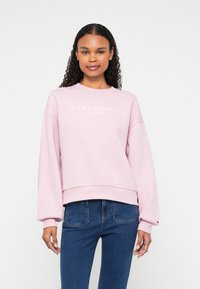 Világos rózsaszín pulóver, oversized fazonnal, bordás mandzsettákkal és szegéllyel, fehér hímzett "TOMMY HILFIGER" logóval. Sötét farmernadrággal párosítva.