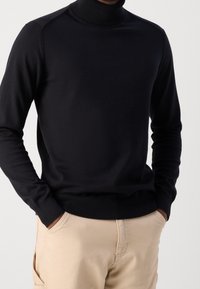 Pull à col roulé noir avec col et poignets côtelés, présentant une coupe ajustée et une texture lisse. Associé à un pantalon beige.