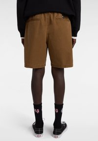 Bruine katoenen shorts met een elastische tailleband, met twee achterzakken. Gecombineerd met zwarte sokken en lage sneakers.