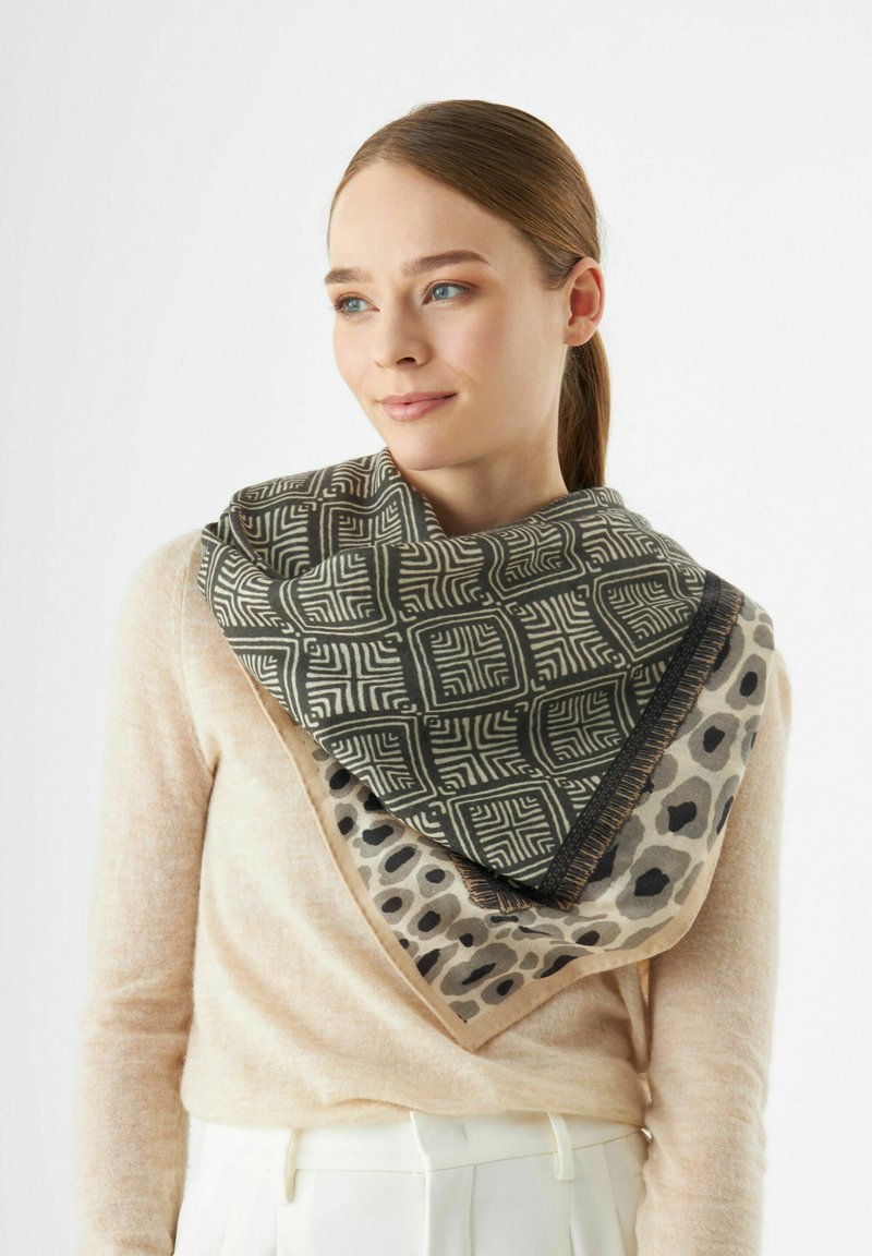 Khaki en beige sjaal met een patroon van geometrische vormen en luipaardprints, met franjes aan de randen, gedragen in een triangulaire drapeerstijl.