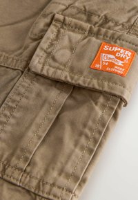 Beige arbetskläder med en iögonfallande orange "Superdry"-etikett. Funktioner inkluderar en ficka, robust sömnad och en mjuk textur.