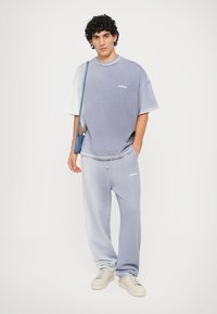 Jonge man staat in een lichtblauw oversized t-shirt en trainingsbroek met witte sneakers en een blauwe schoudertas.