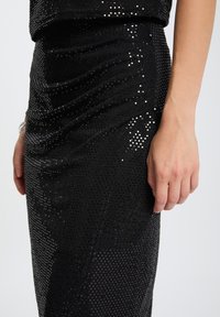 Gonna nera con paillettes e design a bodycon, caratterizzata da un tessuto testurizzato, arricciature e una finitura liscia. Mano appoggiata sulla gonna.
