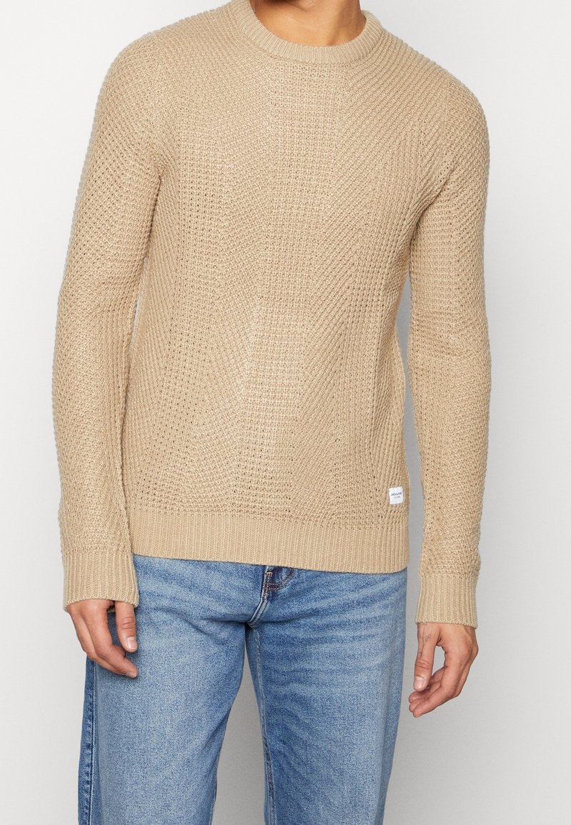 Beige neulepaita, jossa on pyöreä kaula-aukko, ribbireunat hihansuissa ja helmasssa, sekä teksturoitu kuvio. Paita on puettu sinisten farkkujen päälle. Pienet tarrat ala-reunassa.