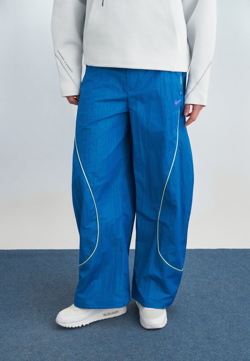 Persona che indossa pantaloni larghi di colore blu acceso con profilatura gialla, sneakers bianche e una maglietta a maniche lunghe grigio chiaro, in piedi su un tappeto grigio.