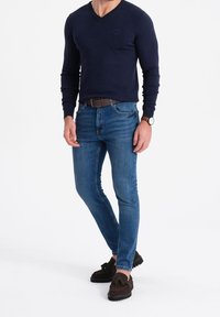 Merimies V-kauluspaita, slim-fit siniset farkut, ruskea vyö ja tummat loafersit tekstuuripinnalla. Yksinkertainen muotoilu ilman näkyvissä olevia kuvioita.