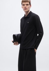 Μαύρο πόλο με μακριά μανίκια και υφή, με ένα μικρό κεντητό λογότυπο. Συνδυάζεται με μαύρο, φαρδύ παντελόνι και clutch.