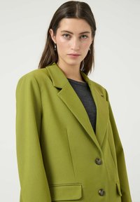 Veste blazer verte cintrée avec revers crantés, deux boutons et poches latérales. Réalisée en tissu texturé, portée avec un haut gris foncé en dessous.