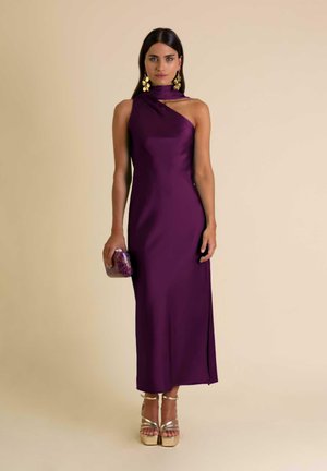 Femme portant une robe asymétrique violette élégante, des talons compensés dorés, tenant une pochette scintillante, sur un fond neutre.