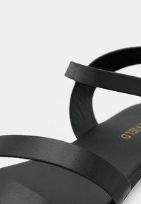 Sandale en cuir noir avec des brides fines et une semelle plate. Texture lisse avec un détail de marque doré sur la semelle intérieure. Design minimaliste.