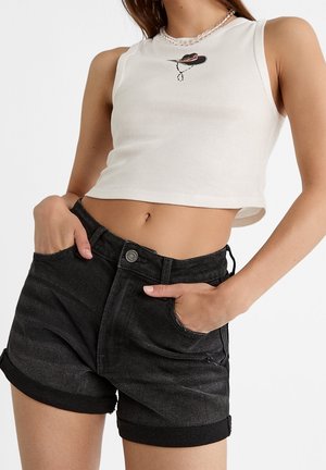 Femme portant un crop top blanc sans manches avec un logo de chapeau et un short en jean noir avec ourlet roulé, mains dans les poches.