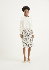 Blusa bianca corta con scollatura arricciata e maniche lunghe, abbinata a una gonna al ginocchio con un motivo di foglie nere su uno sfondo crema.
