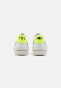 Zapatilla blanca con una suela gruesa, que presenta un logo "Nike" en amarillo brillante y negro en la parte posterior, además de detalles en gris. Textura de cuero suave.