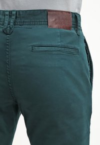 Teal bomullbyxor har en slät textur, framfickor och en brun läderlapp vid midjan med varumärkesprägling.