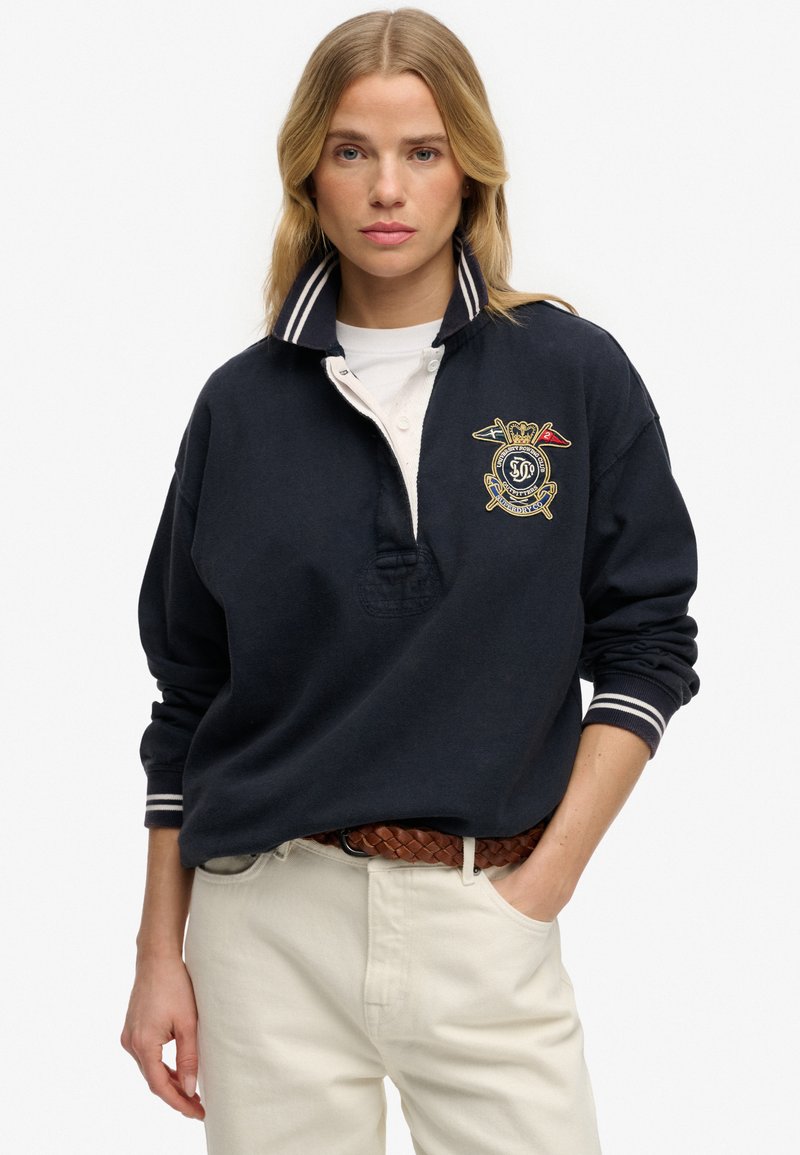 Femme aux cheveux blonds portant un maillot de rugby bleu marine avec un col rayé et un blason brodé, un pantalon blanc et une ceinture tressée marron.