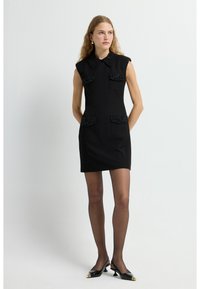 Schwarzes ärmelloses Kleid mit Kragen, bestickten vorderen Taschen und einer figurbetonten Silhouette. Getragen mit transparenten Strumpfhosen und spitzen Absätzen.
