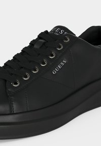 Baskets basses noires avec lacets noirs, œillets argentés et logo "GUESS" sur le côté et la languette, avec une semelle épaisse.
