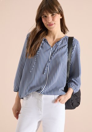 Žena v modro-bílé blůze s vertikálními pruhy a bílých kalhotách, nesoucí černou prošívanou kabelku přes rameno.