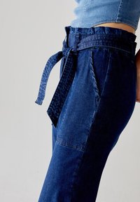 I pantaloni in denim blu scuro presentano una vita arricciata con una cintura in tessuto texture, gamba larga e una tasca laterale. Il tessuto ha una finitura liscia.