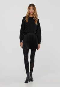 Donna in maglione nero, gonna mini plissettata, collant neri trasparenti con pois bianchi e stivaletti neri con tacco che sta in piedi su uno sfondo bianco.