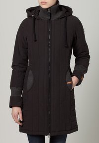 Černý dlouhý péřový kabát s kapucí, vertikální švy, žebrované manžety, dvě velké kapsy a zapínání na zip. Měkká textura a ležérní design.