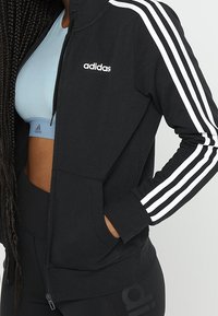 Chaqueta negra con cremallera y diseño de tres rayas blancas en las mangas, que cuenta con un bolsillo frontal y una textura suave. Lleva puesta una camiseta de deporte azul claro debajo.