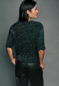 Maglione corto a maniche corte di un verde scuro, con una finitura morbida e pelosa, abbinato a pantaloni neri. Cuciture visibili e un design dalla vestibilità rilassata.
