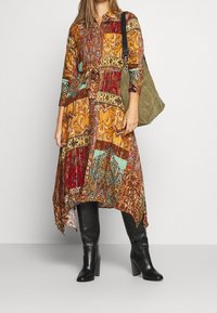 Femme portant une robe à motif patchwork coloré avec des bottes noires à talon montant jusqu'au genou, portant un sac à bandoulière vert olive.