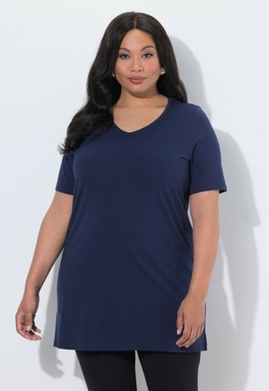 Frau mit langen dunklen Haaren, die ein marineblaues kurzärmeliges Tunika-Top und schwarze Leggings trägt, vor einem schlichten hellen Hintergrund stehend.