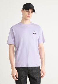 Lilla kortærmet t-shirt lavet af bomuld, med et lille sort trekantet logo på venstre bryst og en afslappet pasform.