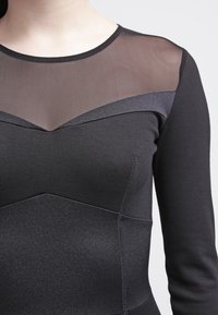 Svart klänning med långa ärmar, en genomskinlig mesh upptill och kontrasterande satin detaljer i en strukturerad design. Slät textur med synliga sömmar.