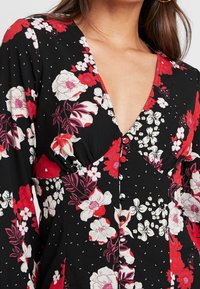 Donna che indossa un abito nero con stampa floreale rossa, bianca e rosa, scollo a V profondo, maniche lunghe e piccoli bottoni coperti di tessuto sul davanti.
