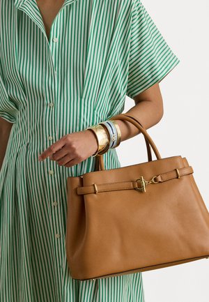 Tan leather handbag with a gold hardware clasp, held with a hand adorned in colorful bangles, paired with a green and white striped shirt dress.
