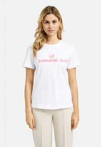 White cotton t-shirt with short sleeves, featuring "SUNSHINE CLUB" in pink and orange text, worn with beige trousers. Simple and casual design.