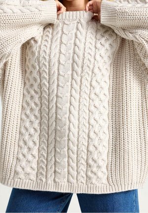 Personne portant un pull oversize crème en maille torsadée, remontant le col, associé à un jean bleu, sur un fond uni.