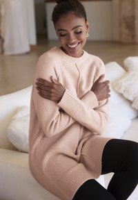 Hellrosa Pullover mit gerippten Bündchen und Saum, im entspannten Schnitt und strukturiertem Strick. Getragen über schwarzen Leggings, sitzend auf einem Sofa.