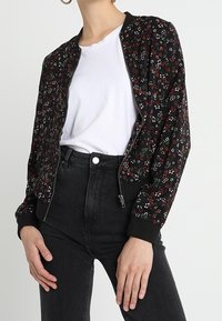 Femme portant une veste zippée noire à motifs floraux sur un t-shirt blanc uni avec un jean noir taille haute, posant avec les mains près des hanches.