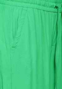 Zielone spodnie z elastycznym pasem i bocznymi kieszeniami, wykonane z miękkiego, lekkiego materiału o gładkiej fakturze i bez widocznych wzorów.