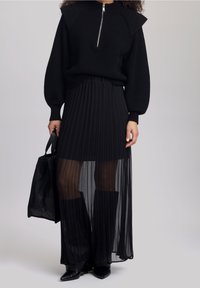 Pull en maille noir avec un col montant et une fermeture éclair, associé à une jupe plissée noire dotée d'un voile transparent. Accessoire : sac noir.