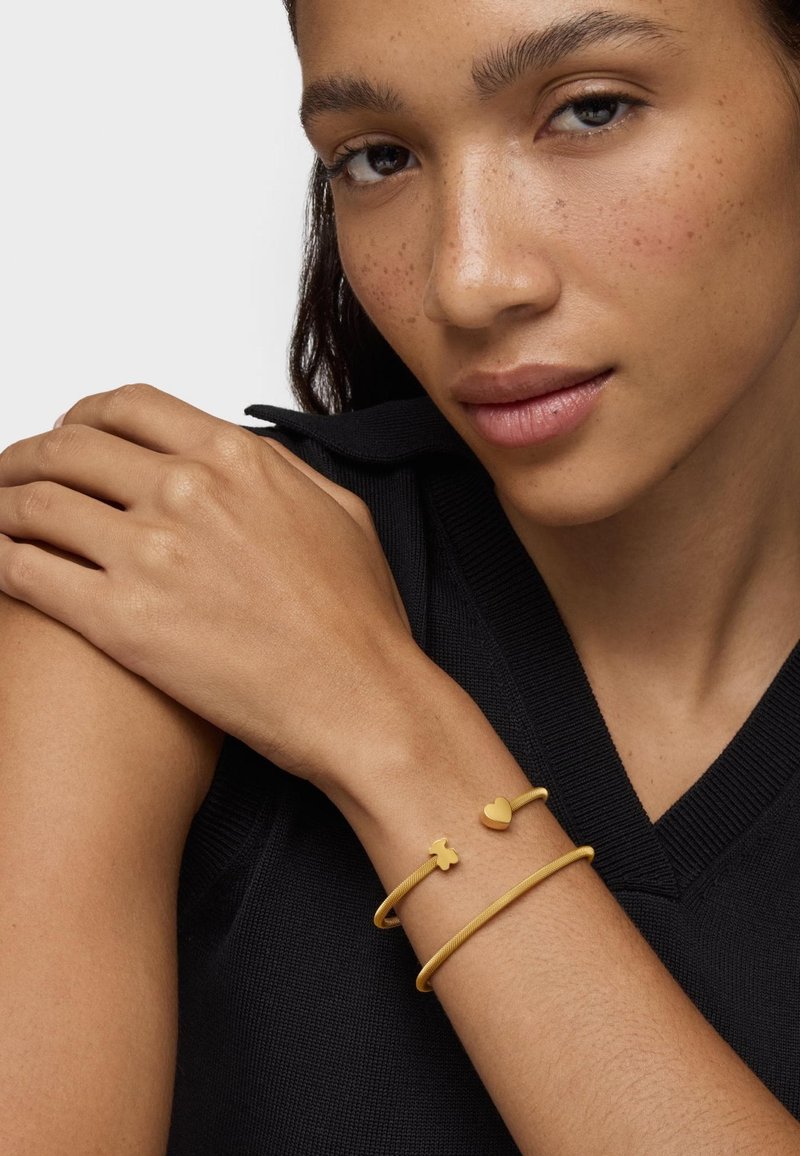 Pulseras de color dorado hechas de metal liso, con formas de corazón y trébol. Llevadas sobre una piel clara, con un suéter negro de fondo.