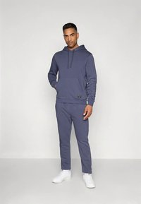 Homem de pé vestindo um conjunto de hoodie e calças de treino azuis com ténis brancos, contra um fundo claro e liso.