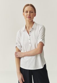 Biała koszulka polo z krótkimi, podwiniętymi rękawami, pięcioma guzikami oraz kołnierzykiem. Materiał wygląda na lekki i lekko teksturowany.