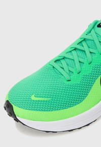 Zapatilla deportiva verde con parte superior de malla texturizada, cordones y un logo negro. Cuenta con una media suela blanca y una suela exterior de goma negra.