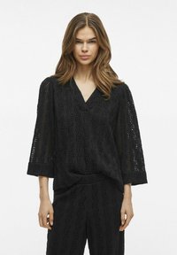 Blusa nera con texture, scollatura a V, maniche 3/4 e dettagli a onda intrecciati; abbinata a pantaloni coordinati con texture.