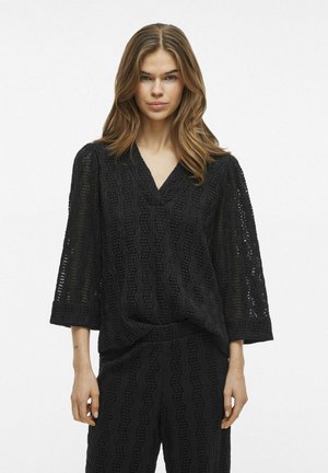 Blusa nera con texture, scollatura a V, maniche 3/4 e dettagli a onda intrecciati; abbinata a pantaloni coordinati con texture.