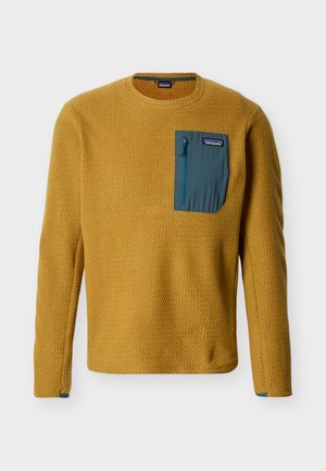 Pikad varrukad, sinepikollane tekstuuriga pullover, millel on tihe sinakasroheline rinna tõmblukuga tasku ja Patagonia logo taskul.